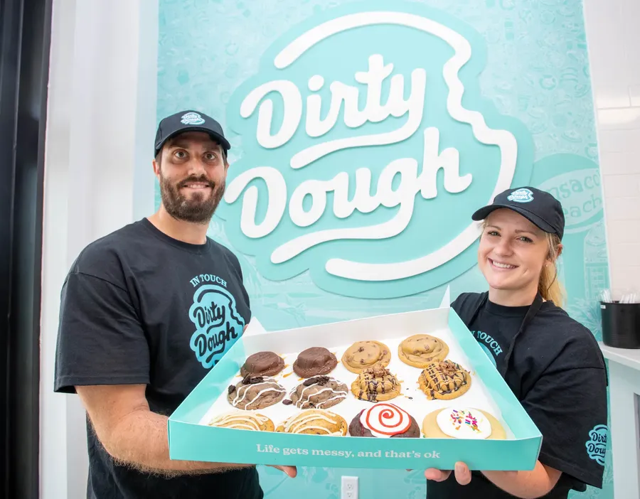 Two Dirty Dough team members holding a box of stuffed cookies in front of the teal Dirty Dough logo wall